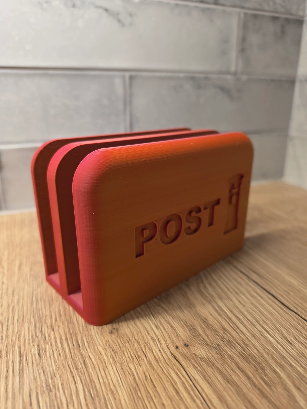 A afterglow 3D-printed post box with post embossed on the front, sitting on a wooden tabletop with a gray tiled wall in the background.