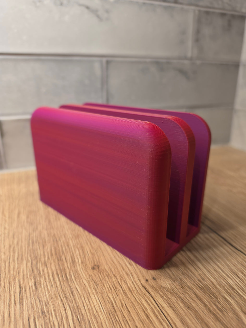 A 3D-printed card holder or coaster stack in a vibrant magenta-to-purple gradient, shown on a wooden tabletop with a gray tiled backsplash in the background.