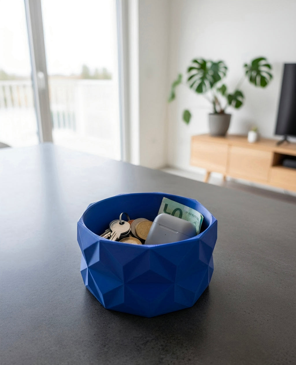 A blue and red geometric Bowl containing keys, coins, and a banknote, sitting on a dark grey tabletop in a modern living room setting.