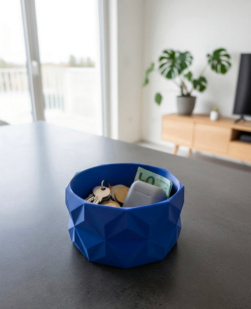 A blue and red geometric Bowl containing keys, coins, and a banknote, sitting on a dark grey tabletop in a modern living room setting.