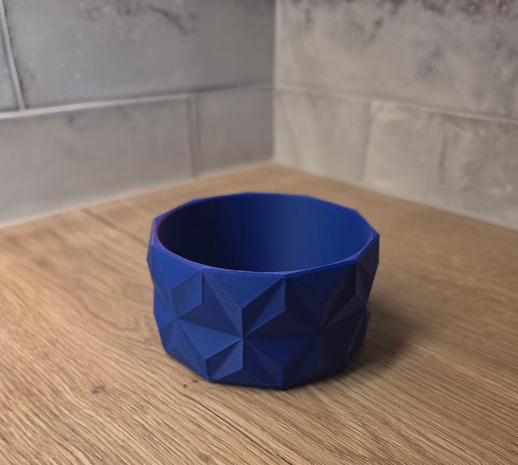A blue and red geometric faceted bowl with a 3D printed texture, shown on a wooden countertop against a tiled backsplash.
