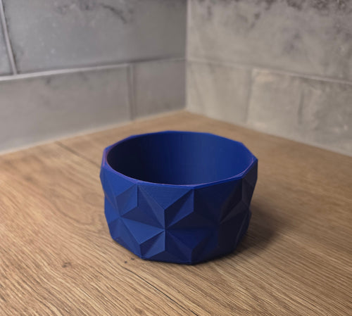 A blue and red geometric faceted bowl with a 3D printed texture, shown on a wooden countertop against a tiled backsplash.