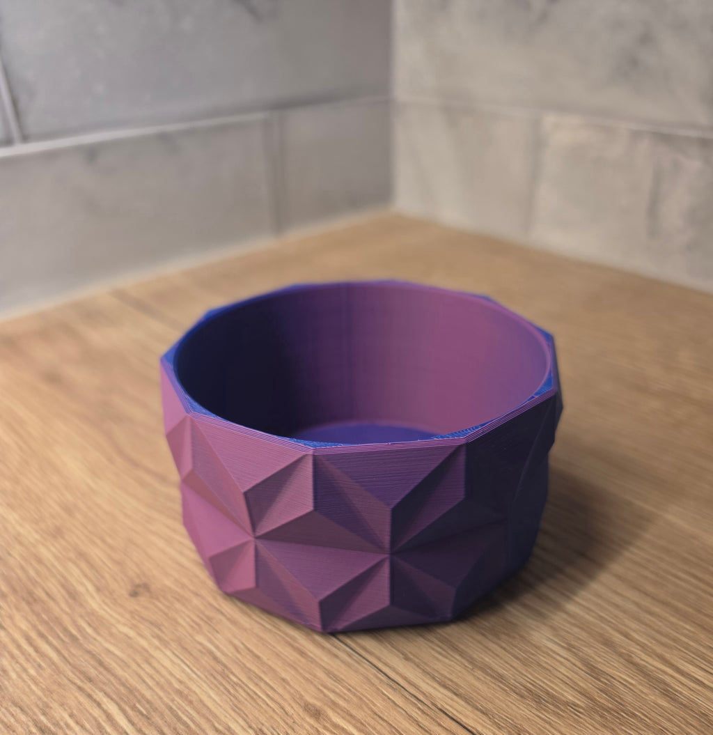 A blue and red geometric faceted bowl with a 3D printed texture, shown on a wooden countertop against a tiled backsplash.
