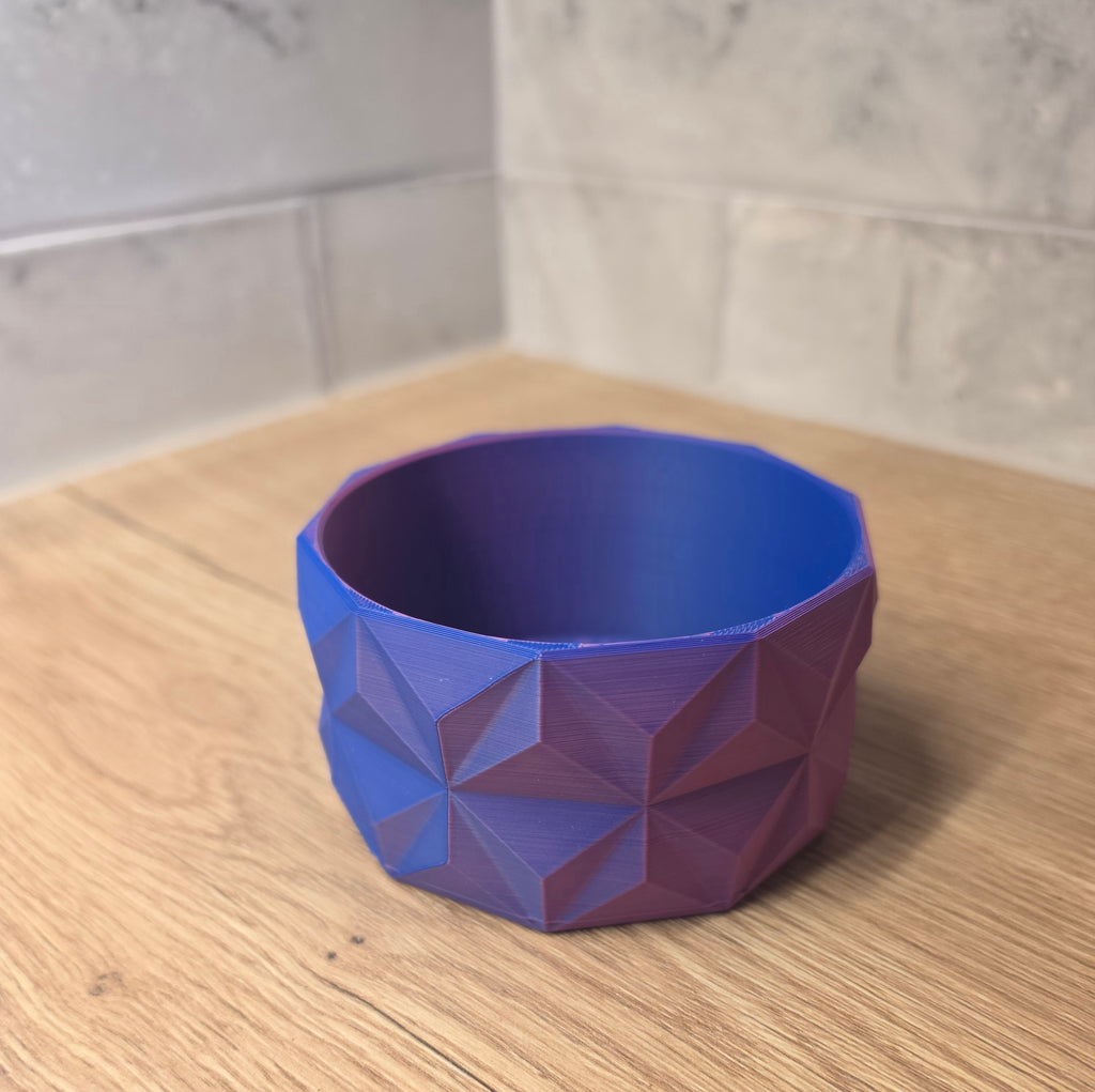A blue and red geometric faceted bowl with a 3D printed texture, shown on a wooden countertop against a tiled backsplash.