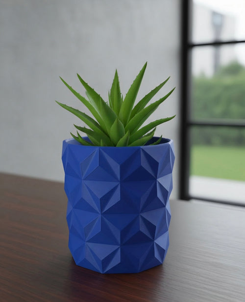 A green succulent plant in a blue /Red faceted geometric planter, placed on a dark wooden table with a blurred window and outdoor view in the background.