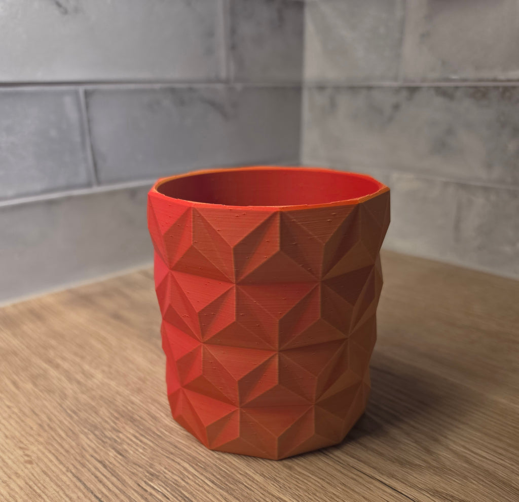 A red and yellow geometric faceted planter standing on a wooden surface against a grey tiled wall.