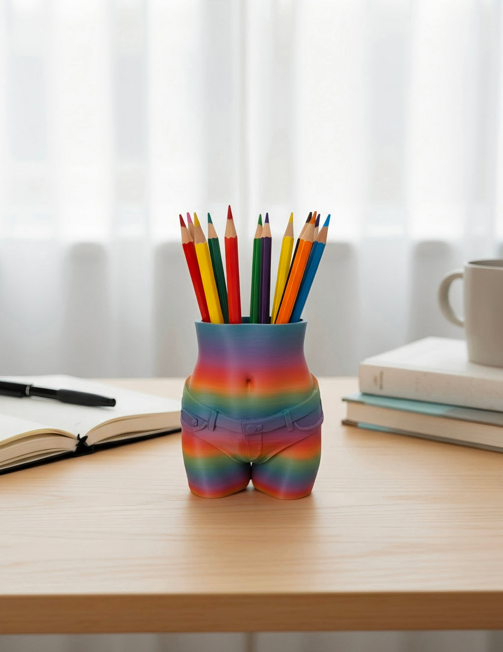 A novelty desk holder shaped like a person's hips and upper thighs, covered in a rainbow gradient pattern, holding several colored pencils. It is placed on a light wood desk with books and a mug in the background.