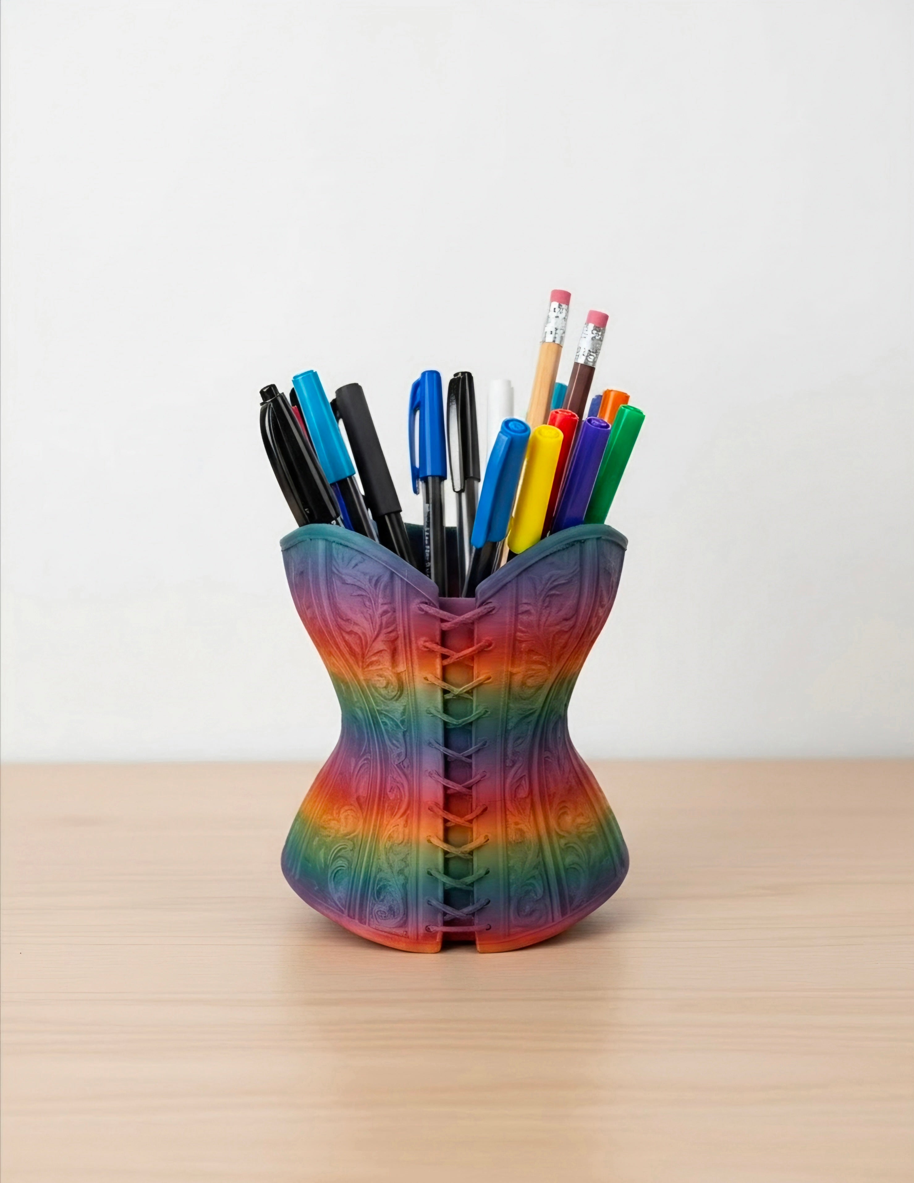 A rainbow-colored pen holder shaped like a corset, filled with various pens and pencils, sitting on a light wood surface against a white wall.