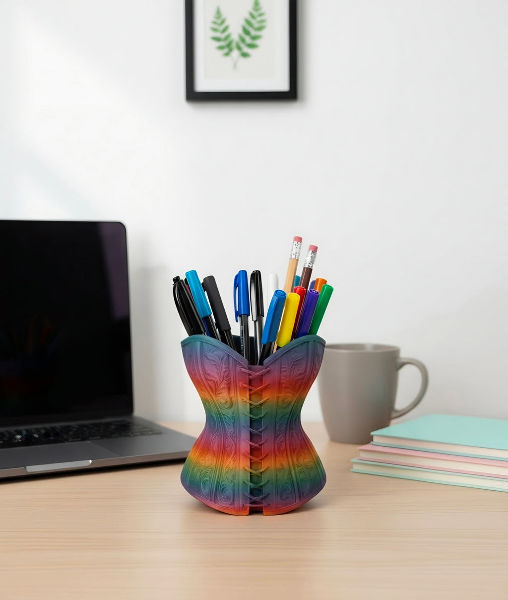 A rainbow pen holder shaped like a corset sits on a light wood desk, filled with pens and pencils, with a laptop, mug, and notebooks in the background.