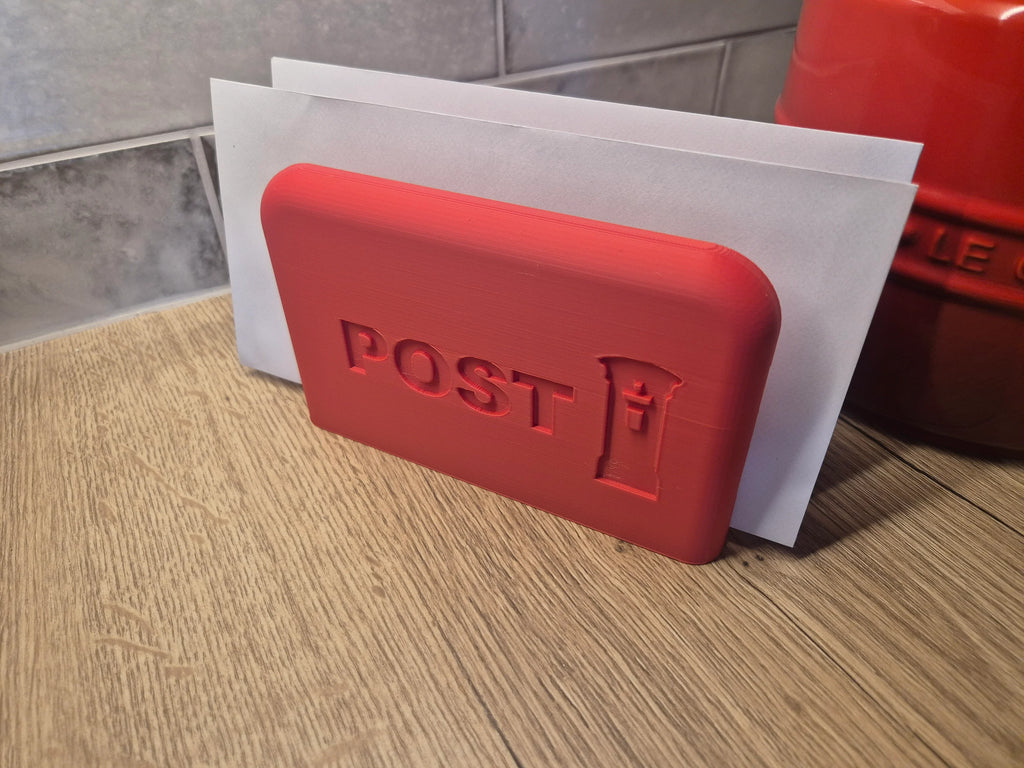A red rectangular postmark-style stamp holder or decorative postcard holder with embossed 'POST' and a small post icon, shown leaning against white envelopes on a wooden surface.