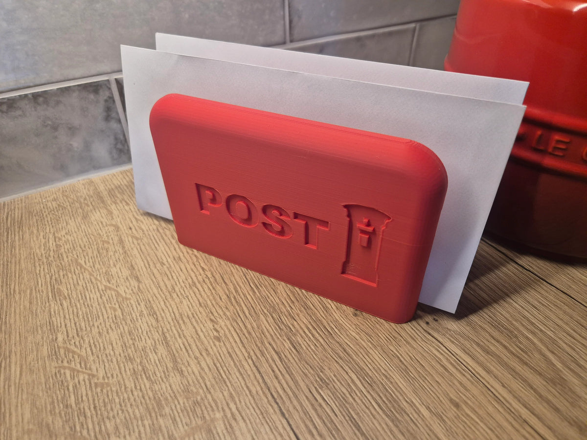 A red rectangular postmark-style stamp holder or decorative postcard holder with embossed 'POST' and a small post icon, shown leaning against white envelopes on a wooden surface.
