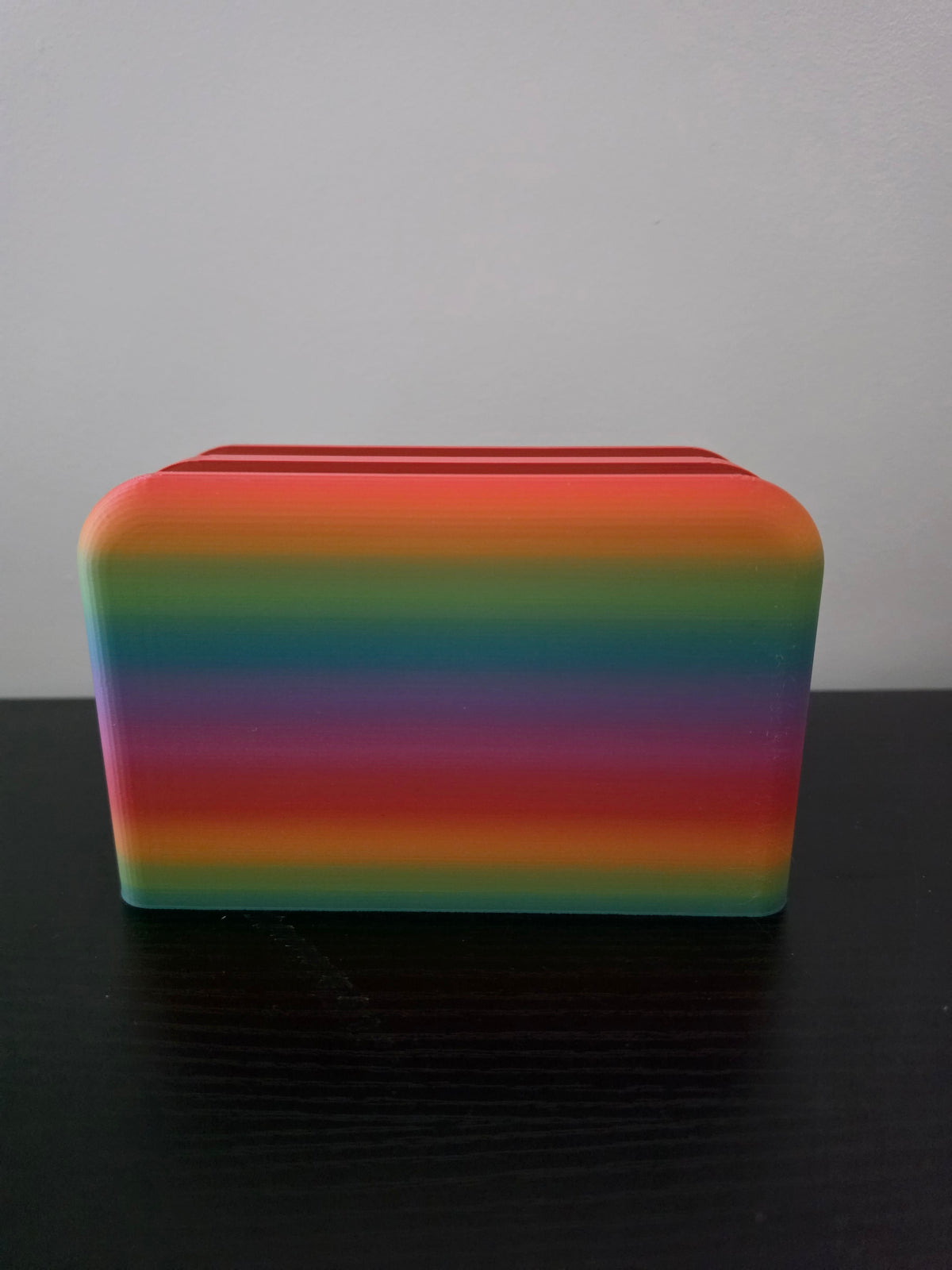 A rectangular rainbow-striped object with rounded corners sits on a dark surface against a light gray background.