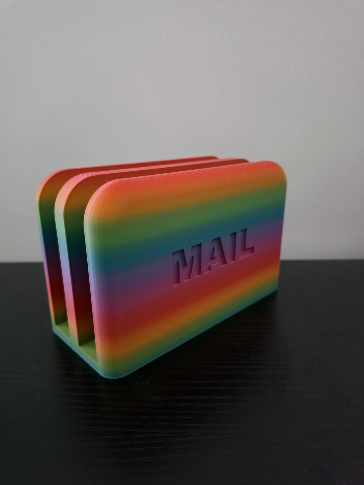 A 3D-printed rainbow-striped desk organizer with the word 'MAIL' embossed on the front, standing on a dark surface against a light gray background.