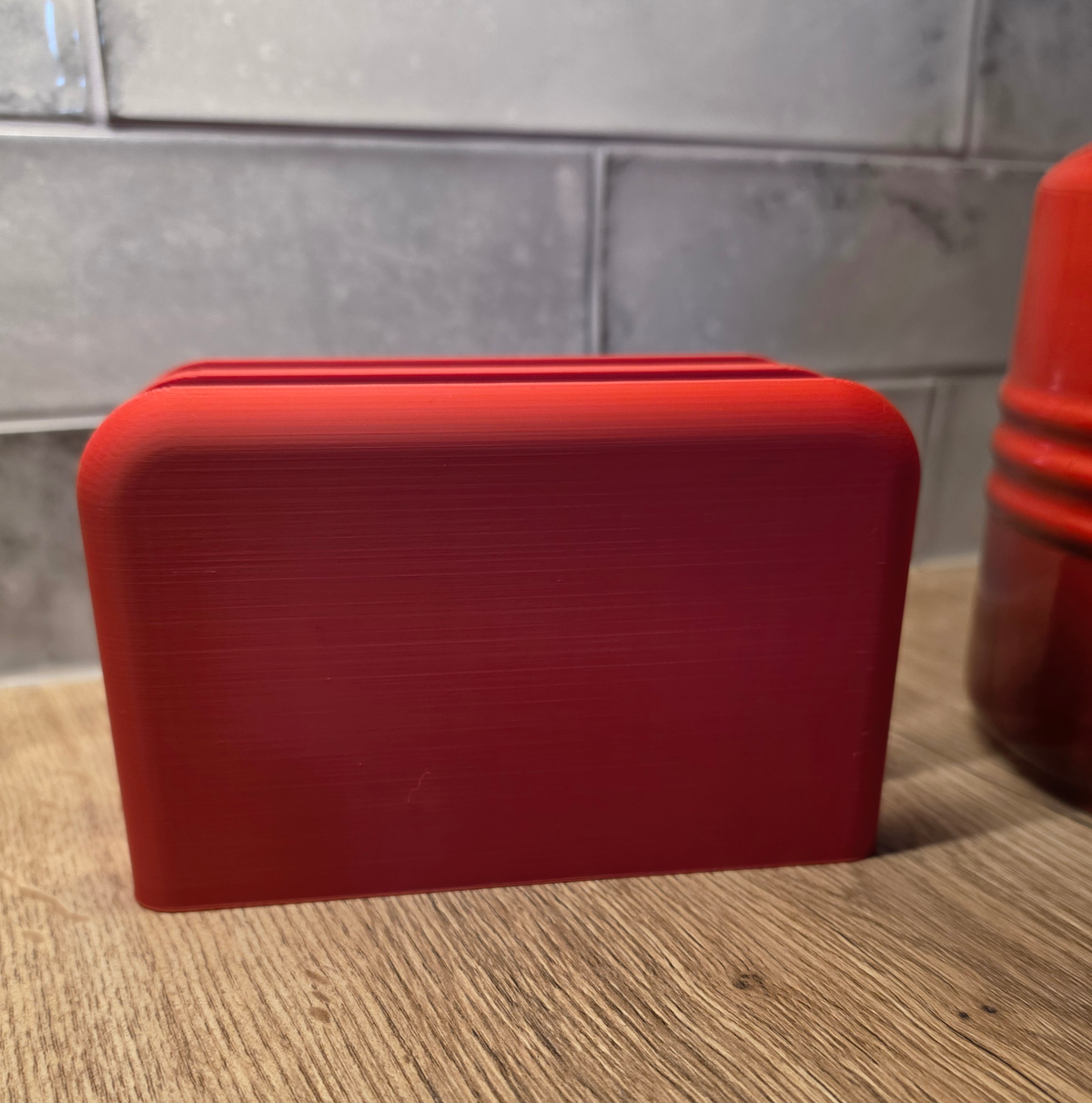 A bright red rectangular 3D-printed object with rounded corners and horizontal slot lines on top, shown on a wooden surface against a gray tiled wall.