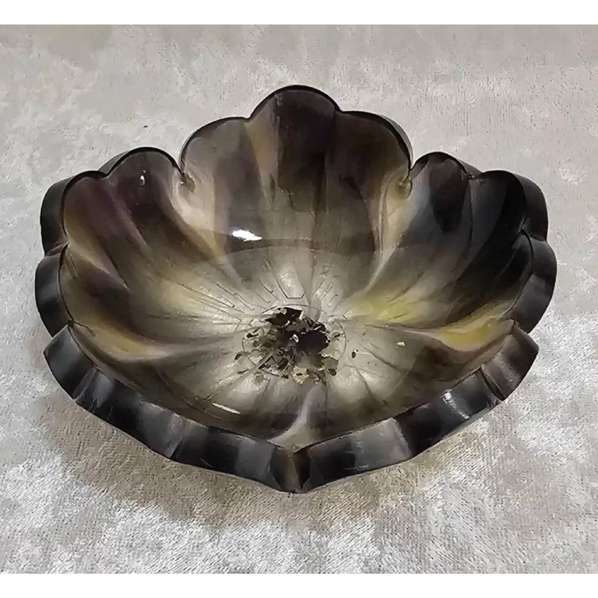 A translucent petal-shaped glass bowl with a scalloped rim, featuring a smoky gradient from dark gray and black at the edges to a lighter gray and subtle yellow tint near the center, resting on a textured light gray surface.