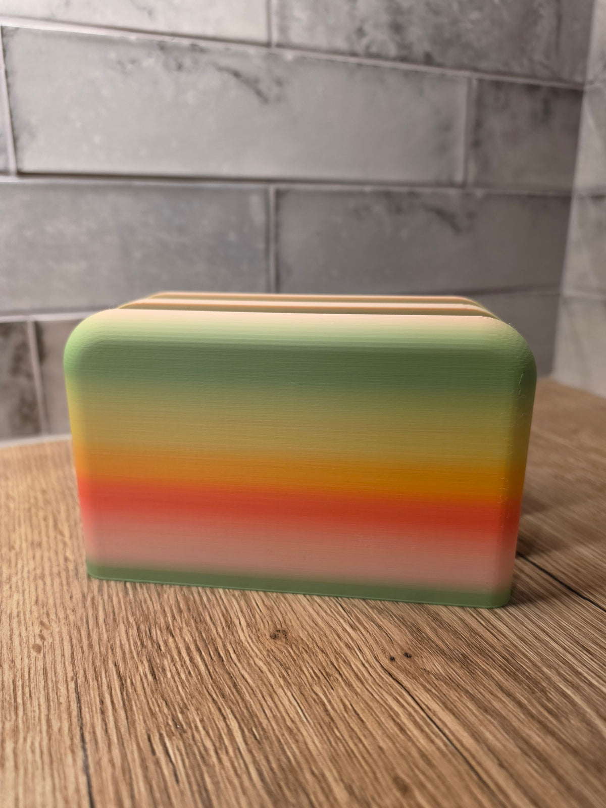 A rectangular container with rounded corners, featuring a horizontal striped gradient design in green, yellow, orange, and white, sitting on a wooden surface with a gray tiled background.