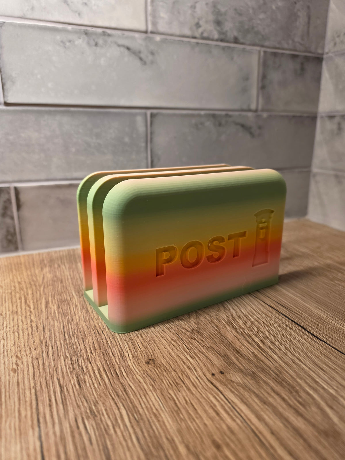 A rectangular desk organizer with rounded corners, featuring a green-to-orange gradient design and the embossed word 'POST' on the front, set on a wooden tabletop against a gray tiled wall.