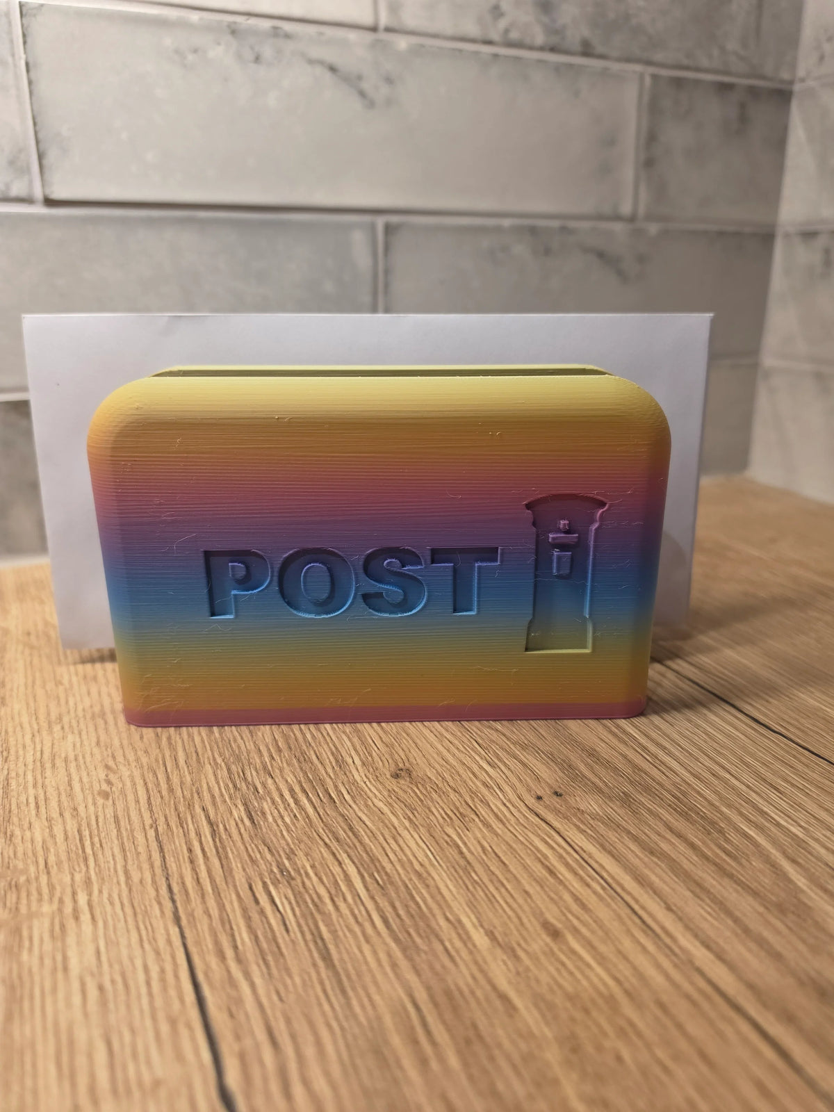 A 3D-printed rainbow-colored post box with the word 'POST' and a small post icon embossed on the front, placed on a wooden surface with a tiled wall in the background.
