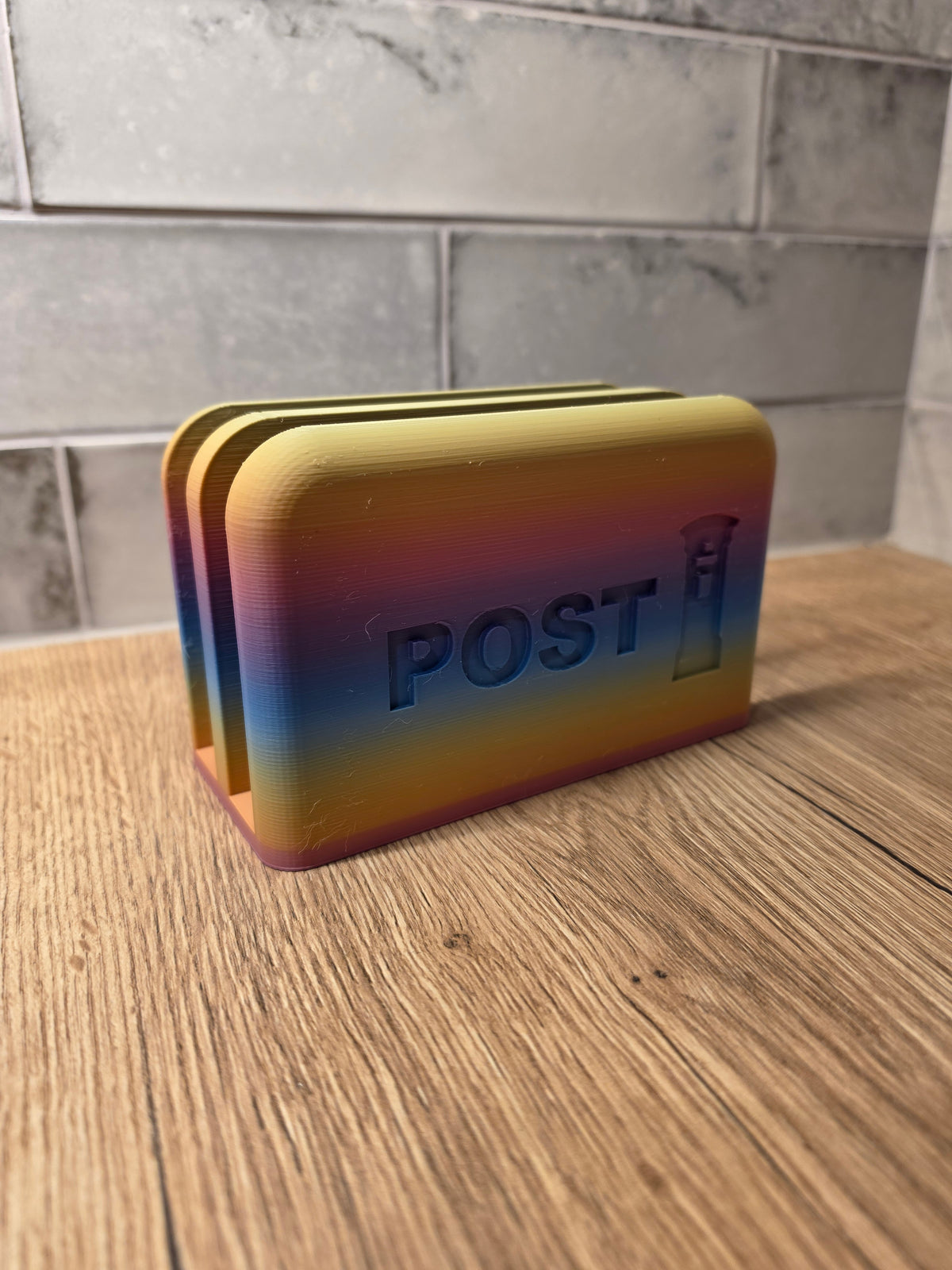 A 3D-printed post box with a vibrant rainbow gradient finish, featuring the word 'POST' and a small envelope icon on the front, set on a wooden surface against a gray tiled background.