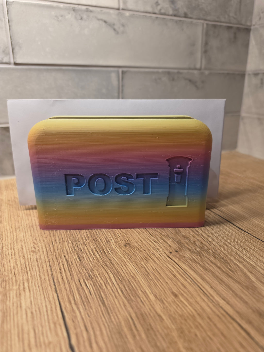 A 3D-printed rainbow-colored post box with the word 'POST' and a small post icon embossed on the front, placed on a wooden surface with a tiled wall in the background.